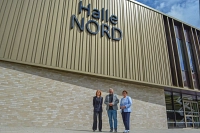 Drei Personen stehen vor einer riesigen Sporthalle mit der Aufschrift Halle Nord.